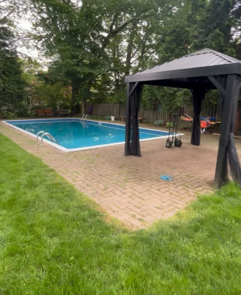 Backyard swimming pool with gazebo and paver patio surrounded by lush greenery.